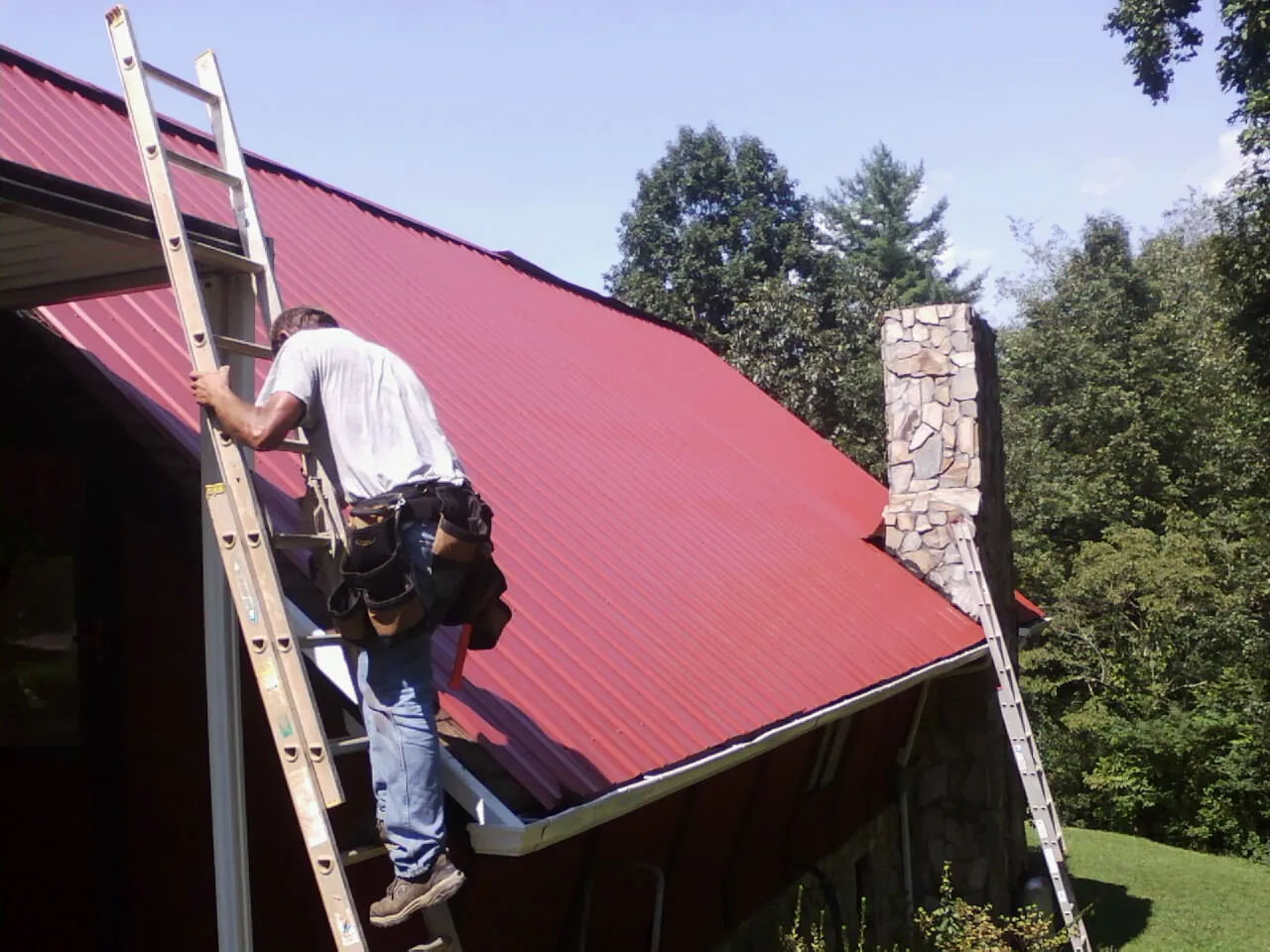 Roofer on a ladder beside a red metal roof and stone chimney — Soffit & Fascia Repair by Roofing Brothers in Zwingle